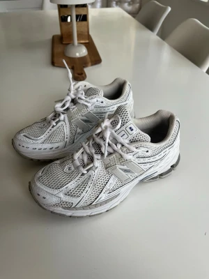 Vita New Balance sneakers mesh Bra Skick! - Säljer ett par vita New Balance sneakers med klassisk mesh och syntetpaneler. Skorna har en sportig look med grå detaljer och den ikoniska N-loggan på sidan. Perfekta för dig som gillar retrostil och bekväma skor till vardags.