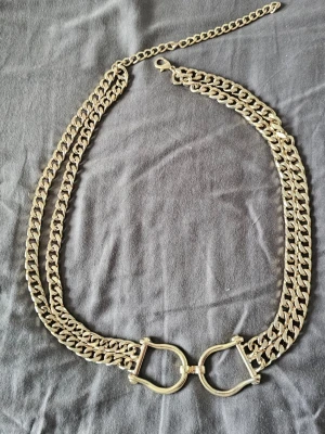 Oanvänt vintage kedjebälte i guld med hästskospänne (New Old Stock) - Som midjebälte (över klänning/kavaj): Passar storlek M och L (eftersom det kan ställas in mellan 79 cm och 98 cm).Som höftbälte (lågt sittande): Passar storlek S och M. Riktigt vintage plagg, aldrig använd. Perfekt för vintagestyle!
