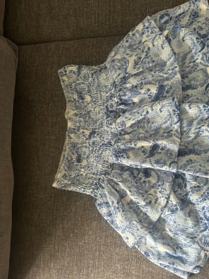 Blåvit kortkjol från Gina Tricot - Supersöt blåvit kortkjol från Gina Tricot med volang och smock i midjan. Kjolen har mönster och är gjord i ett lätt och luftigt material, perfekt för varma dagar. Passar dig som gillar en feminin och trendig stil.