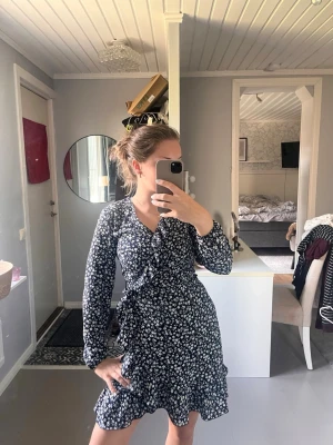 Blommig omlottklänning i mörkblått - Supersöt mörkblå kortklänning med vitt blommönster och omlottdesign. Klänningen har långa ärmar, volangkant nertill och markerad midja med knyt i sidan. Perfekt för dig som gillar en feminin och trendig stil.