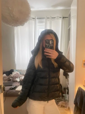 Marinblå pufferjacka med huva - Säljer en marinblå pufferjacka med huva och dragkedja framtill. Jackan har en klassisk quiltad design och är vadderad för extra värme. Perfekt för kalla dagar och har en stilren look som passar till det mesta.