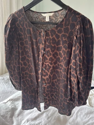 Leopardmönstrad blus från H&M - Säljer en brun och svart leopardmönstrad blus med knappar framtill och rund halsringning, i linne-material. Använd endast en gång. Köpt förra sommaren. 