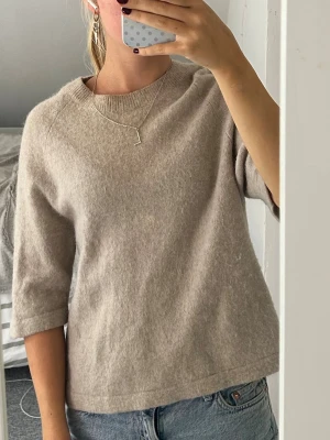 Beige stickad tröja med trekvartsärm - Mysig beige stickad tröja med trekvartsärm och rund hals. Tröjan har en avslappnad passform. Perfekt att matcha med jeans eller kjol för en enkel och stilren look.