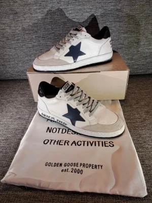 Golden Goose vita sneakers med Stjärna och Text - Golden Goose sneakers i vitt läder med grå mockadetaljer, svart hälparti och en mörkblå stjärna på sidan. Skorna har grå skosnören och textdetaljer på sidan och hälen. Klassisk låg modell med retrovibbar och slitna detaljer. Snabb Leverans!