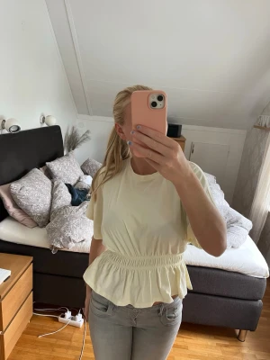 Ljusgul topp med volang och resår - Säljer en ljusgul topp med korta ärmar och rund hals. Toppen har resår i midjan som ger en snygg volang nedtill och en feminin siluett. Perfekt till jeans eller kjol för en fräsch look.