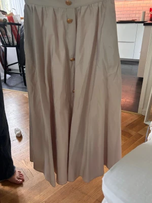 Beige långkjol med knappar - Säljer en beige långkjol med hög midja och dekorativa knappar framtill. Kjolen har en lätt utsvängd passform och är tillverkad i ett mjukt, tunt material som ger ett snyggt fall. Perfekt för dig som gillar stilrena och enkla plagg. Litet hål vid ena fickan, knappt märkbart.
