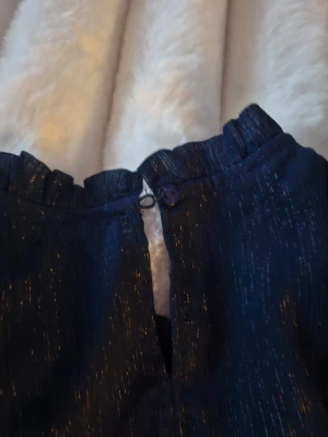 Mörkblå glittrig blus med volang från gina tricot  - Säljer en mörkblå blus med glittriga detaljer och volang vid halsen. Klänningen har lång ärm och knäppning bak i nacken. Perfekt för dig som vill sticka ut lite extra med en elegant look.