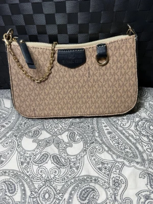 Beige handväska  - Snygg beige handväska med klassiskt MK-mönster över hela väskan. Väskan har guldfärgade detaljer, kedjehandtag och svarta läderdetaljer. Perfekt storlek för det viktigaste och stängs med dragkedja.