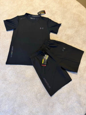 Svart träningsset från Under Armour - Säljer ett svart träningsset från Under Armour med t-shirt och shorts. Setet är gjort i ett lätt och snabbtorkande funktionsmaterial, perfekt för löpning eller gym. Diskreta loggor på både tröja och shorts samt elastisk midja på shortsen.