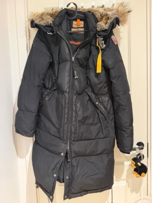 Svart lång vinterjacka med päls, parajumper - Snygg svart lång dunjacka från Parajumpers med avtagbar huva och fuskpäls. Jackan har dragkedja, tryckknappar, flera fickor och den klassiska gula remmen vid halsen. Perfekt för kalla vinterdagar och riktigt varm.