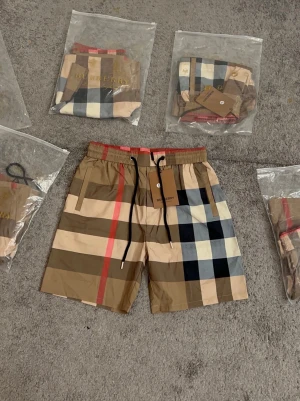 Burberry shorts  - Säljer ett par klassiska Burberry shorts i den ikoniska rutiga designen med beige, svart, röd och vit färg. Shortsen har elastisk midja med svart snörning och är tillverkade i ett lätt syntetmaterial, perfekta för sommaren.