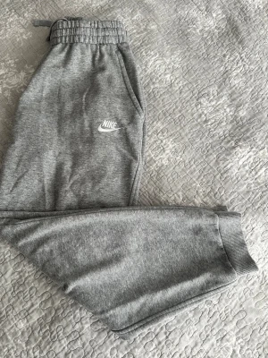 Gråa mjukisbyxor från Nike XL ( 158/170 cm) - Snygga grå mjukisbyxor från Nike med resår i midjan och muddar vid bensluten. Byxorna har sidofickor och en broderad Nike-logga på låret. Tillverkade i mjukt och bekvämt material, perfekta för chill dagar eller träning.
