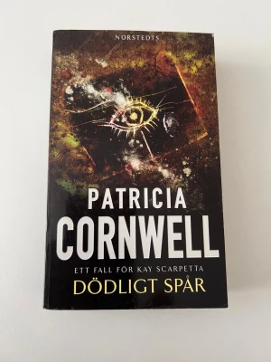Dödligt spår av Patricia Cornwell - Spännande kriminalroman av Patricia Cornwell, del i serien om Kay Scarpetta. Boken har ett mörkt omslag med ett mystiskt öga i centrum och är utgiven av Norstedts. Perfekt för dig som gillar deckare och thrillers med starka karaktärer och oväntade vändningar. Några sidor i boken har vikts sedan tidigare. 