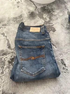 Blå jeans från Nudie Jeans - Säljer ett par klassiska blå jeans från Nudie Jeans med raka ben och snygga slitningar. Jeansen har fem fickor och den ikoniska broderade vågen på bakfickan. Tillverkade i slitstarkt denim med orangea sömmar och patch i läder bak.