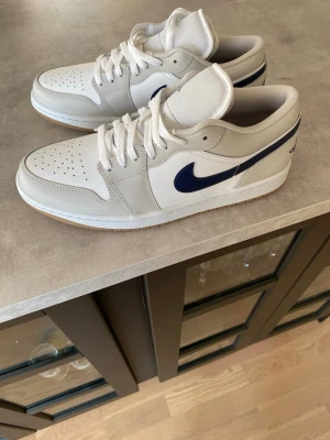 Vita och blå Nike Air Jordan 1 Low (helt nya) - Säljer ett par fräscha Nike Air Jordan 1 Low sneakers i vitt och marinblått. Skorna har klassisk låg siluett, perforerad tåbox och vit sula med gummiyttersula. Materialet är en mix av läder och textil, med den ikoniska Swoosh-loggan i blått på sidan.               white/midnight navy/neutral grey/med brown är exakta färgen
