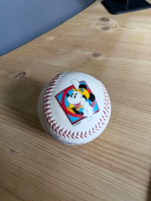 Baseboll med Musse Pigg-motiv - Cool baseboll i vitt med röda sömmar och ett färgglatt tryck av Musse Pigg i basebollutstyrsel. Ena sidan har Musse Pigg-logga och den andra sidan har texten 'Mickey Mouse' i blått. Perfekt för samlare eller Disney-fans.