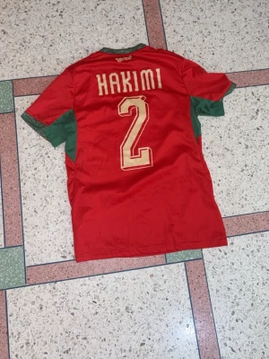 Marocko fotbollströja Hakimi #2 Puma - Säljer en röd Marocko fotbollströja från Puma med Hakimi och nummer 2 på ryggen. Tröjan har gröna detaljer på ärmar och sidor, samt officiellt landslagsemblem på bröstet. Tillverkad i funktionsmaterial Drycell som andas bra. Finns även andra storlek