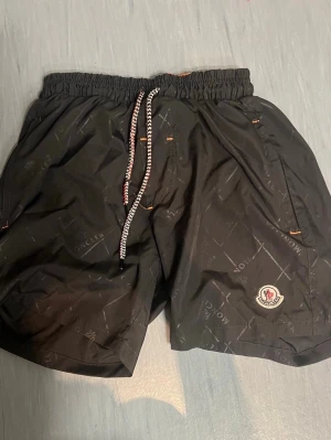 Svarta Moncler shorts med snörning - Säljer ett par svarta shorts från Moncler med diskret mönster och logga på benet. Shortsen har elastisk midja med snörning och sidofickor. Perfekta för sommaren och har en sportig vibe.