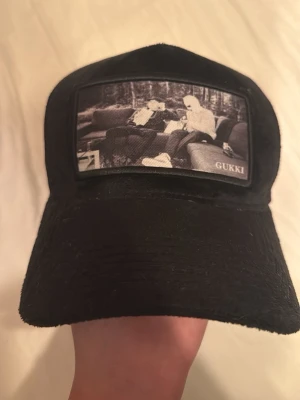 Svart keps från Gukki Clothing - Snygg svart keps från Gukki Clothing med mesh på baksidan och ett svartvitt tryck framtill som visar två personer på en soffa. Kepsen har böjd skärm och justerbar passform. Perfekt för dig som gillar streetwear och vill sticka ut. Nästan aldrig använt och riktig bra skick