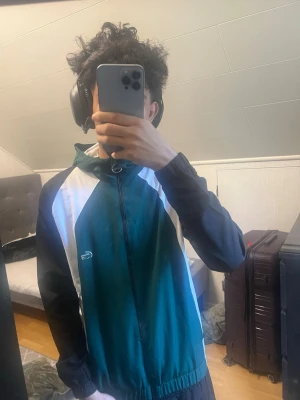 Grön och vit vindjacka från Lacoste - Snygg vindjacka från Lacoste i grönt, vitt och svart. Jackan har dragkedja framtill, huva och elastiska muddar. Klassisk Lacoste-logga på bröstet och stor text på ryggen. Perfekt för dig som gillar sportig och retro stil.