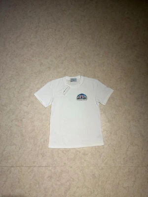 Vit t-shirt från Casablanca - Säljer en vit t-shirt från Casablanca med ett färgglatt tryck och logga på bröstet. T-shirten har rund halsringning och korta ärmar. Tillverkad i mjukt bomullsmaterial och har en clean, stilren look med exklusiv känsla.
