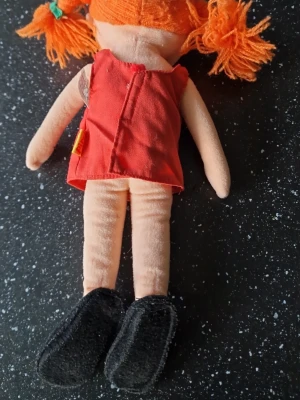 Pippi Långstrump mjukdocka - Söt mjuk docka med orange garnhår i tofsar, röd klänning med grön ficka och svarta tygskor. Dockan har broderat ansikte och är gjord i mjukt tyg, perfekt som kramkompis eller dekoration.