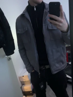 Grå mockajacka med dragkedja - Snygg grå mockajacka med raka linjer och två bröstfickor med lock. Jackan har hel dragkedja framtill och klassisk krage. Perfekt för dig som vill ha en stilren och modern look. Passar till det mesta och är enkel att matcha.