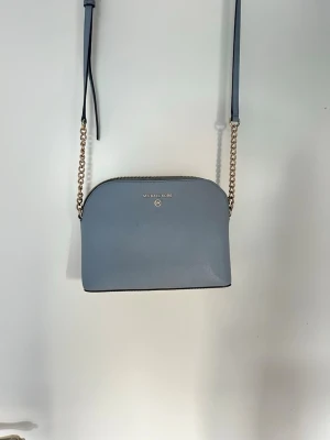 Ljusblå axelväska Michael Kors - Snygg ljusblå axelväska från Michael Kors i slätt skinn. Väskan har guldfärgade kedjedetaljer på axelremmen och dragkedja upptill. Insidan är fodrad med logotypmönster och har en innerficka med dragkedja. Perfekt storlek för det viktigaste.