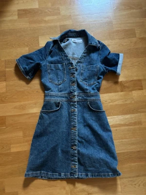 Jeansklänning med knappar från ZARA - Snygg jeansklänning från ZARA med korta ärmar, krage och knappar framtill. Två bröstfickor och markerad midja ger en cool och avslappnad look. Perfekt till sneakers eller boots för en trendig outfit.