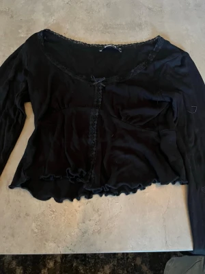 Svart topp med spets från Gina Tricot - Säljer en svart långärmad topp från Gina Tricot i storlek XS. Toppen har spetsdetaljer vid halsringningen och framtill, liten rosett och vågig kant nertill. Materialet är mjukt och stretchigt, perfekt för en trendig look.