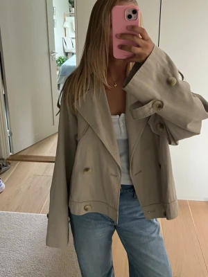Beige cropped trenchcoat - Trendig beige cropped trenchcoat med oversized passform, stora slag och dubbelknäppning. Jackan har breda ärmar med dekorativa knappar och är tillverkad i ett tunt, mjukt tyg som ger en avslappnad look. Perfekt för lager på lager under vår och höst.