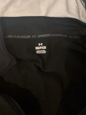 Svart träningströja från Under Armour - Svart långärmad träningströja från Under Armour med halv dragkedja framtill och hög krage. Tröjan har en vit logga på bröstet och är tillverkad i ett mjukt, stretchigt material som passar perfekt för träning och sportaktiviteter.