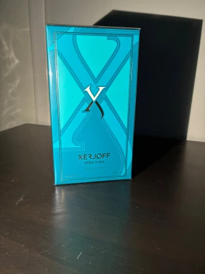 Xerjoff Erba Pura Eau de Parfum - Xerjoff Erba Pura Eau de Parfum i en snygg turkosblå förpackning med gulddetaljer. Lådan har en modern design med ett stort X på framsidan och elegant text. Parfymen är känd för sin fruktiga och fräscha doftprofil och passar dig som gillar exklusiva dofter.