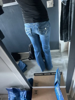 Blå bootcut jeans med broderade fickor - Snygga blå bootcut jeans med broderade detaljer på bakfickorna och klassisk femficksdesign. Jeansen har en ljus tvätt och subtila slitningar framtill. Perfekta för dig som gillar en avslappnad men trendig stil.