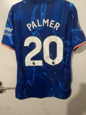 Chelsea FC blå matchtröja Palmer 20 - Säljer en blå Chelsea FC matchtröja från Nike med Palmer och nummer 20 på ryggen. Tröjan har ett unikt mönster i olika blå nyanser, korta ärmar och klubbmärke på bröstet. Perfekt för dig som älskar fotboll och vill stötta ditt lag.