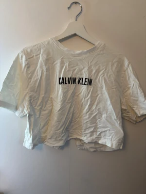 Vit croppad t-shirt Calvin Klein - Snygg vit croppad t-shirt från Calvin Klein med svart logotyptryck på bröstet. T-shirten har korta ärmar och är tillverkad i mjuk bomull, perfekt för en avslappnad och trendig look.