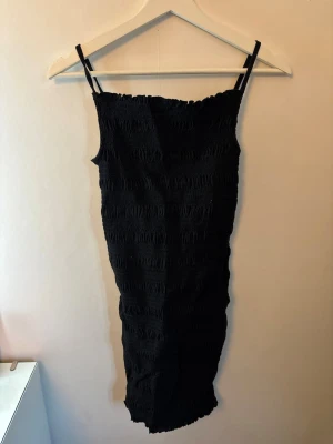 Svart klänning - Svart klänning med smockad struktur och tunna axelband. Klänningen har en kroppsnära passform och är ärmlös, vilket ger en stilren och enkel look. Perfekt för dig som gillar minimalistisk stil och vill ha något bekvämt men ändå snyggt.