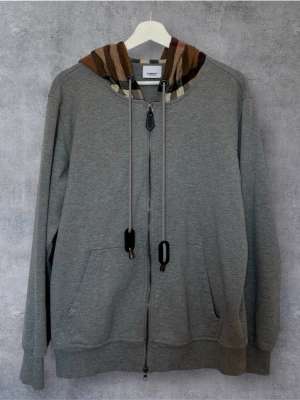 Burberry Zip Hoodie  - Elegant och stilren zip hoodie från Burberry i klassisk grå färg. Modellen har den ikoniska Burberry-rutiga huvan, två framfickor samt högkvalitativ dragkedja framtill. Tillverkad i mjuk och bekväm bomullsblandning med premiumkänsla och tidlös design som passar året runt.  Skick: Mycket fint skick Färg: Grå Detaljer: Burberry Check-huva, fullzip, två framfickor, justerbar huva Passform: Regular fit  Passar S,M liten L