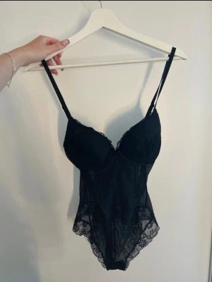 Svart spetsbody med tunna axelband - Snygg svart body i spets med tunna, justerbara axelband och vadderade kupor. Bodyn har genomskinliga spetsdetaljer och vacker kant i spets runt ben och byst. Perfekt för dig som gillar en feminin och elegant look.