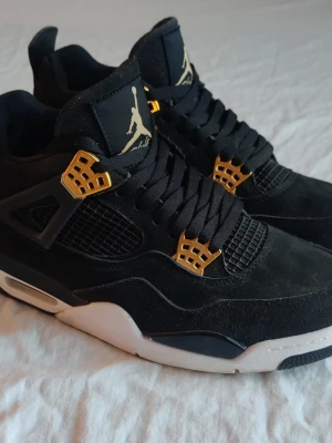 Svarta Jordan 4 med gulddetaljer - Stilrena svarta sneakers med gulddetaljer och vit sula. Ovandelen är i mocka och mesh, med svarta skosnören och coola guldaccenter vid snörningen. Perfekta för dig som vill sticka ut med en klassisk men ändå unik look.