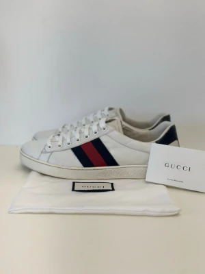Vita Gucci ave sneakers - Säljer ett par klassiska vita Gucci sneakers i läder med de ikoniska blå och röda ränderna på sidan. Skorna har vita snören, låg profil och mörkblå detalj vid hälen. Perfekta för dig som gillar stilrena och exklusiva sneakers.