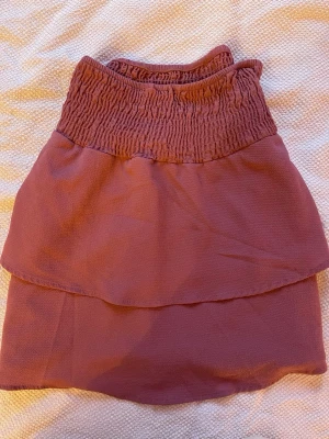 Rosa volangkjol med smock - Söt rosa kjol med bred smock i midjan och dubbla volanger. Kjolen har en mjuk och luftig känsla, perfekt för dig som gillar en feminin och avslappnad stil. Passar till både sneakers och sandaler.