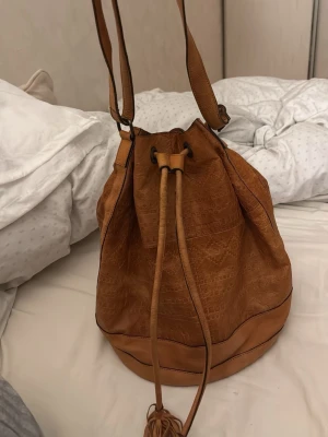 Brun bucket bag i läderimitation - Snygg brun bucket bag med dragskostängning och axelrem. Väskan har en slät yta med krokopräglat mönster och dekorativa tofsar. Perfekt för dig som vill ha plats för allt du behöver under dagen. Insidan är rymlig och har en mörk textilfoder.
