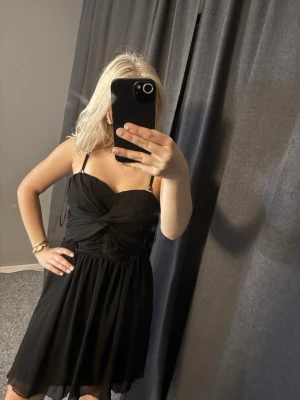 Svart klänning från Bik Bok - Säljer en svart klänning från Bik Bok med smala axelband och twistad detalj framtill. Klänningen har en lätt utsvängd kjol och dragkedja i sidan. Perfekt för dig som gillar en enkel men snygg look.