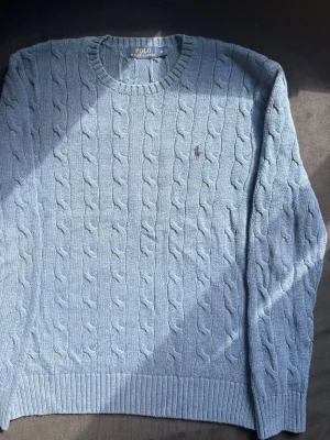Ljusblå kabelstickad tröja Polo Ralph Lauren - Kabelstickad ljusblå tröja från Polo Ralph Lauren med rund halsringning och klassisk broderad logga på bröstet. Tröjan har ribbade muddar vid ärmslut och nederkant. Perfekt för dig som gillar stilrena och tidlösa plagg.
