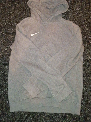 Grå hoodie från Nike med huva - Säljer en stilren grå hoodie från Nike med klassisk huva och vit Nike-logga på bröstet. Hoodien har långa ärmar, ribbade muddar och en avslappnad passform. Perfekt för en chill och sportig look.