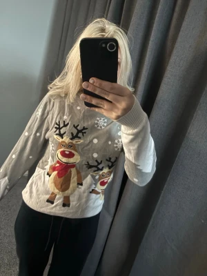 Grå jultröja med renmotiv - Grå stickad jultröja från Christmas Sweats med gulligt renmotiv och snöflingor framtill. Tröjan har långa ärmar och ribbade muddar vid ärmslut och hals. Perfekt för att maxa julkänslan med ett lekfullt print.