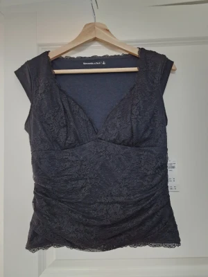 Mörk spets topp med v-ringning - Snygg mörk topp med v-ringning och spetsdetaljer. Ärmlös modell med figurnära passform och fina spetskanter både upptill och nedtill. Perfekt för dig som vill ha en elegant och stilren look.