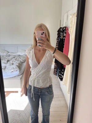 Vit blus - Säljer en vit omlottblus med knytband i midjan och djup v-ringning. Blusen har korta ärmar och fina broderade detaljer vid axlarna. Man kan knyta den både baktill eller framtill. 