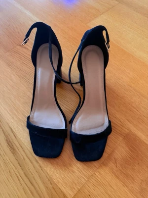 Svarta pumps med blockklack. Aldrig använt - Snygga svarta pumps med bred blockklack och öppen tå. Skorna har en fyrkantig tå, rem över foten och justerbar ankelrem med guldfärgat spänne. Materialet ser ut att vara mockaimitation som ger en lyxig känsla.